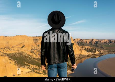 Vista posteriore dell'uomo in un cappello nero e una giacca in pelle con una fotocamera rétro mentre osserva un canyon mozzafiato con un fiume tortuoso sotto un clea Foto Stock