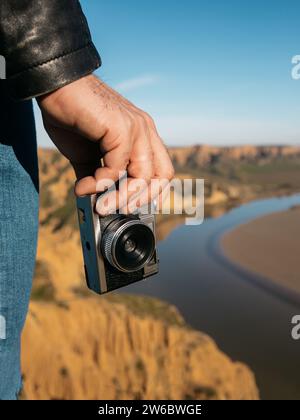 Primo piano della mano di un viaggiatore che tiene in mano una macchina fotografica vecchio stile con uno scenografico paesaggio fluviale sullo sfondo Foto Stock