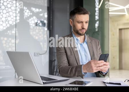 Uomo d'affari maturo, serio e concentrato, che pensa di leggere i social network online, uomo maturo esperto che tiene il telefono utilizzando uno smartphone sul posto di lavoro all'interno dell'ufficio con un computer portatile. Foto Stock