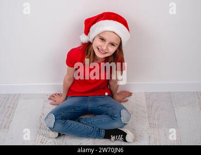 Little Girl con T-Shirt rossa e Santa Hat si siede sul pavimento, per Una foto natalizia su sfondo bianco Foto Stock