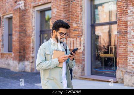 Un giovane studente indiano in tuta e una camicia verde sta camminando fuori da un campus universitario con uno zaino e un notebook e sta usando un telefono cellulare. Foto Stock
