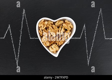 Vista dall'alto delle noci sbucciate in un recipiente a forma di cuore su un piatto di ardesia nera con cardiogramma a battito cardiaco disegnato con gesso Foto Stock