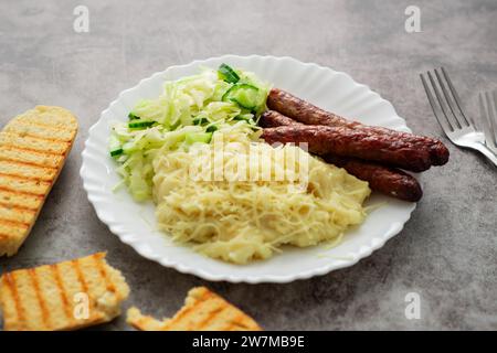 Un piatto appetitoso con purè di patate con formaggio, una rinfrescante insalata di cavoli e cetrioli e succulente salsicce, il tutto splendidamente presente Foto Stock
