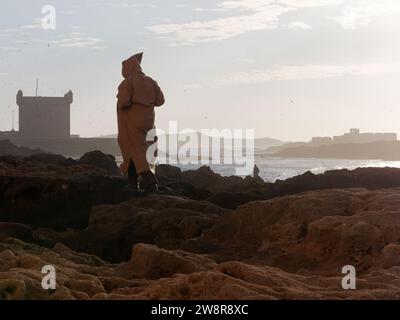 Uomo che indossa una djellaba sulle formazioni rocciose con una torre e un mare alle spalle al tramonto a Essaouira, "la città ventosa", Marocco. 21 dicembre 2023 Foto Stock