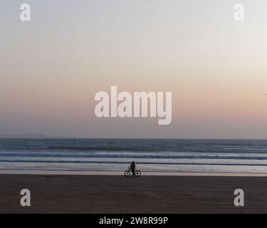 Ciclista solitario in silhouette su una spiaggia al tramonto mentre le onde si infrangono, a Essaouira, "la città ventosa", in Marocco. 21 dicembre 2023 Foto Stock