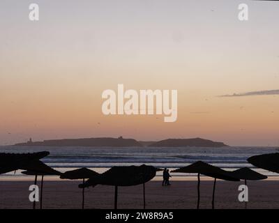 Una coppia cammina su una spiaggia sabbiosa con ombrelloni al tramonto con un'isola alle spalle, a Essaouira, la "città ventosa", in Marocco. 21 dicembre 2023 Foto Stock