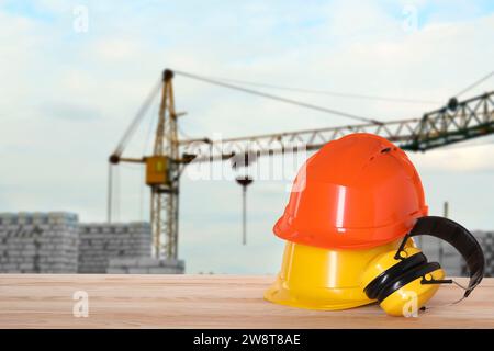 Attrezzatura di sicurezza. Cappelli rigidi e cuffie protettive su superfici in legno vicino al cantiere con gru a torre all'aperto, spazio per testo Foto Stock