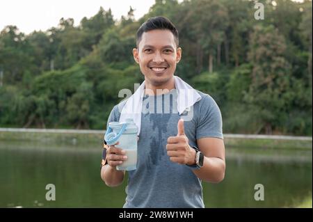 Un uomo asiatico felice, forte e in forma con abbigliamento sportivo sta mostrando il pollice e bevendo un frullato proteico dopo una lunga corsa all'aperto, bevendo una bevanda sportiva Foto Stock