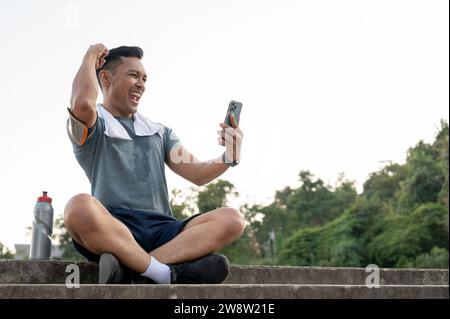 Un allegro uomo asiatico in abbigliamento sportivo riposa sulle scale dopo essersi allenato all'aperto, mostrando il suo pugno, trionfo, guardando lo schermo del suo telefono con una h Foto Stock