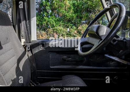 "Sedile del conducente e volante in tessuto nero di una Land Rover Defender; attraverso il finestrino è visibile la foresta." Foto Stock