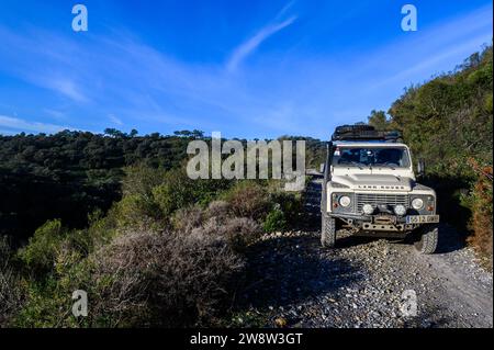 White Land Rover Defender equipaggiata per 4x4 e viaggi via terra, con verricello, su una strada sterrata grigia nel mezzo della macchia, sotto il cielo blu Foto Stock