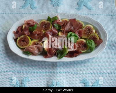 Orizzontalmente direttamente dall'alto, ripresa ravvicinata di insalata di fichi e prosciutto adornata con foglie di basilico fresco servita su un delicato piatto bianco su un tradizionale Foto Stock