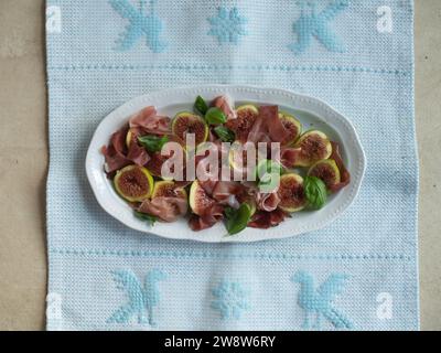 Orizzontalmente direttamente dall'alto, ripresa ravvicinata di insalata di fichi e prosciutto adornata con foglie di basilico fresco servita su un delicato piatto bianco su un tradizionale Foto Stock