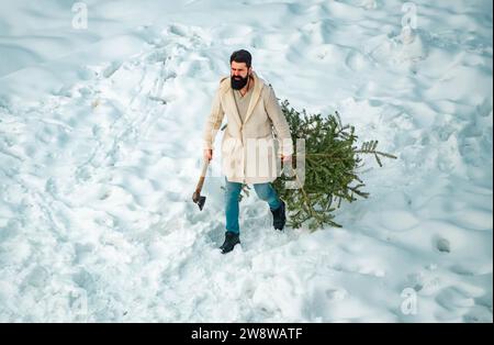 L'uomo barbuto porta l'albero di Natale nel bosco. Taglio dell'albero di Natale. Buon Natale e buone feste. Giovani taglialegna orso abete nel Foto Stock