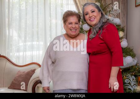 In vita, ritratto frontale orizzontale di due eleganti donne latinoamericane mature, albero di Natale su sfondo sfocato, abito rosso formale, sorriso felice Foto Stock