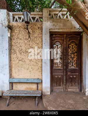 Panca blu in legno intemprato si trova invitante accanto ad una classica porta grunge decorata in legno. Una lampada in metallo d'epoca è appesa sopra la panca, per aggiungere un tocco nostalgico alla scena Foto Stock