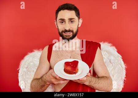 felice uomo barbuto in costume di cupido con una deliziosa torta a forma di cuore e guardando la macchina fotografica sul rosso Foto Stock
