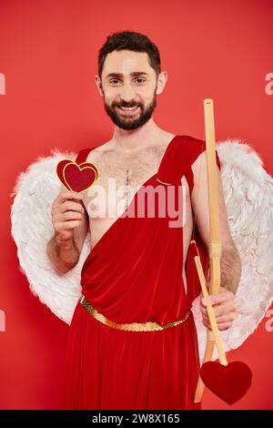 felice uomo barbuto in costume di cupido con cuori di carta e arco con freccia sul rosso, giorno dei san valentini Foto Stock