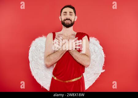 felice uomo barbuto in costume di cupido in piedi con le mani sul petto e guardando la fotocamera sul rosso Foto Stock
