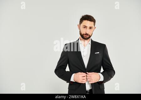 bellissimo uomo d'affari barbuto che abbraccia un elegante abito nero e guarda la fotocamera su sfondo grigio Foto Stock