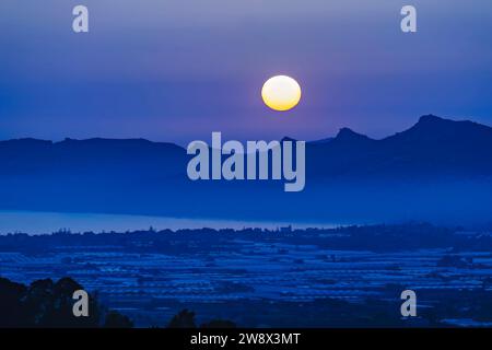 Luna piena che si innalza sopra le tranquille montagne blu e le vette sagomate. Luna piena sul tranquillo paesaggio montano. Foto Stock
