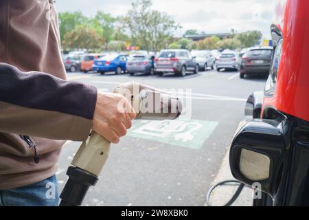 Tenere a mano un caricabatterie per auto elettriche presso un parcheggio con il cartello di ricarica EV a terra. Auto non riconoscibili parcheggiate sullo sfondo. Foto Stock