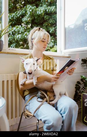 Donna bionda con cane Bull terrier che legge rivista sulla sedia in sala d'attesa all'ospedale animale Foto Stock