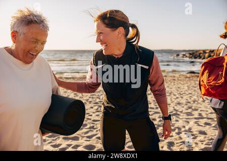 Una bella coach che ride con una donna anziana che tiene il tappetino in spiaggia Foto Stock