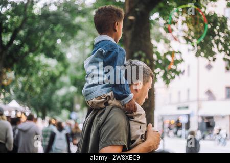 Vista laterale del ragazzo seduto sulla spalla del padre mentre fa shopping al mercatino delle pulci Foto Stock