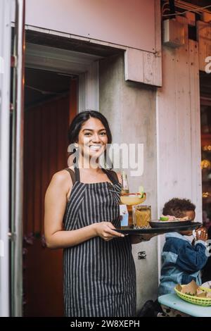 Ritratto di una cameriera sorridente che indossa un grembiule con vassoio di cibo e bevande al bar Foto Stock