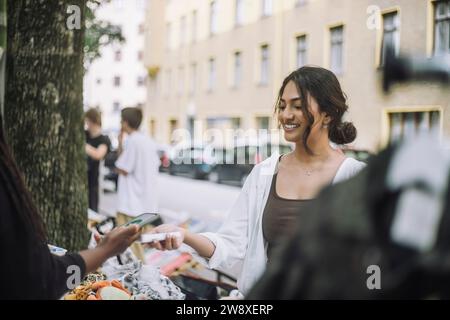 Proprietaria sorridente che prende il pagamento mobile da un cliente che fa acquisti al mercato Foto Stock