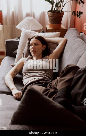 Giovane donna con la mano alzata che guarda lontano mentre si sdraiava sul divano nell'appartamento Foto Stock