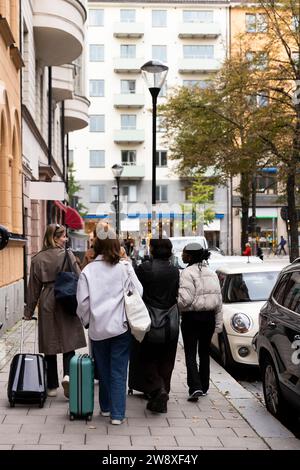 Vista posteriore delle giovani amiche turistiche che camminano sul marciapiede con i bagagli in città Foto Stock