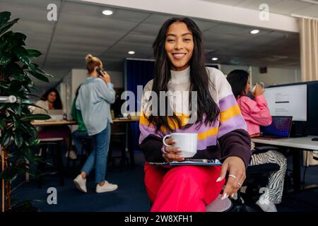 Ritratto di una donna d'affari sorridente che regge un bicchiere mentre è seduta in ufficio Foto Stock