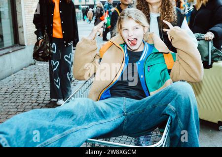 Ragazza che sporge la lingua e il segnale del corno con gesti seduto sul carrello della spesa al sentiero Foto Stock