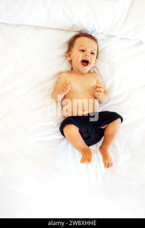 Il bambino simpatico sta ridendo e sdraiato su un lenzuolo bianco sul divano. Un bambino sta sorridendo sul letto a casa. Dolce e felice bambino si sta divertendo. Visualizza Foto Stock