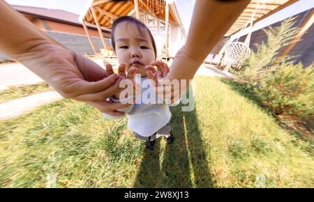 Primo piano delle mani di un genitore e di una graziosa bambina asiatica. Dolce bambino coreano all'aperto su un prato con erba verde. Mamma e bambino stanno camminando nel bac Foto Stock