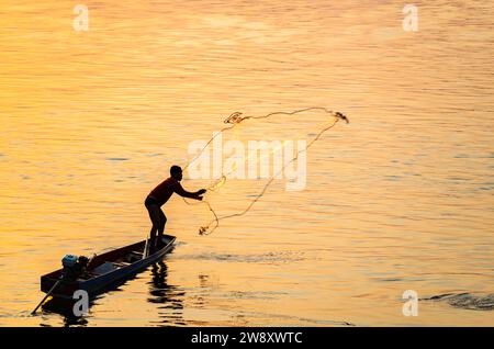 Silhouette di una figura umana che lancia una piccola rete da pesca dalla sua stretta barca, vicino al tramonto, sulle acque del fiume vicino all'isola Don Det, parte del si Foto Stock