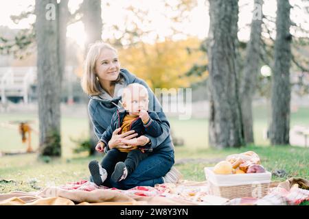 La madre sorridente con un bambino in braccio si siede su un picnic su una coperta accanto a un cestino di cibo Foto Stock