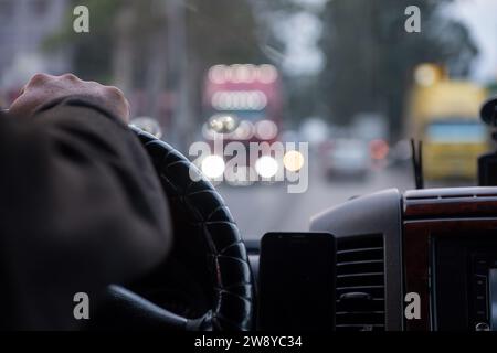 le mani di un vecchio mentre si guida su strada con luci sfocate e traffico automobilistico Foto Stock