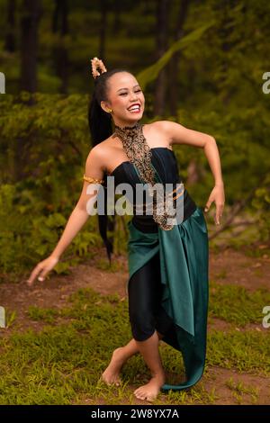 Donna indonesiana con lunghi capelli neri che indossa un costume verde con trucco davanti alla foresta durante il festival Foto Stock