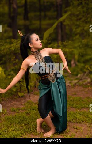 Donna indonesiana con lunghi capelli neri che indossa un costume verde con trucco davanti alla foresta durante il festival Foto Stock