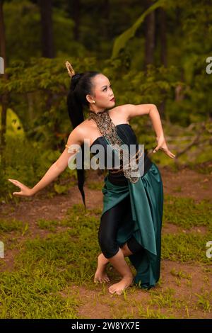 Donna indonesiana con lunghi capelli neri che indossa un costume verde con trucco davanti alla foresta durante il festival Foto Stock