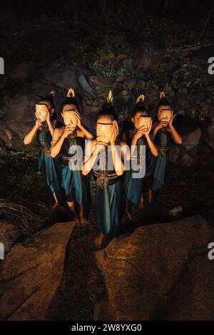 Un gruppo di donne asiatiche che indossano maschere di legno in costumi verdi mentre si trovano tra le rocce all'interno della foresta durante la notte Foto Stock