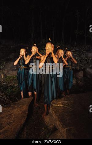 Un gruppo di donne asiatiche che indossano maschere di legno in costumi verdi mentre si trovano tra le rocce all'interno della foresta durante la notte Foto Stock