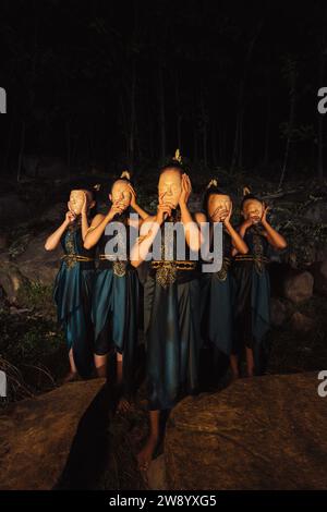 Un gruppo di donne asiatiche che indossano maschere di legno in costumi verdi mentre si trovano tra le rocce all'interno della foresta durante la notte Foto Stock