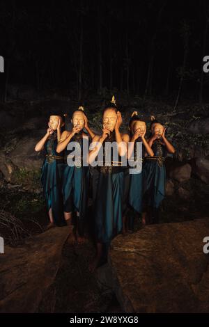 Un gruppo di donne asiatiche che indossano maschere di legno in costumi verdi mentre si trovano tra le rocce all'interno della foresta durante la notte Foto Stock