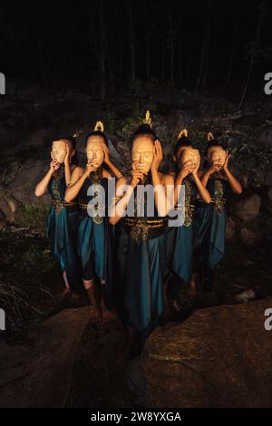 Un gruppo di donne asiatiche che indossano maschere di legno in costumi verdi mentre si trovano tra le rocce all'interno della foresta durante la notte Foto Stock