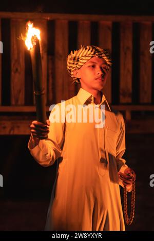 Un uomo arabo che tiene una torcia di fuoco davanti a una casa di legno nella notte buia Foto Stock