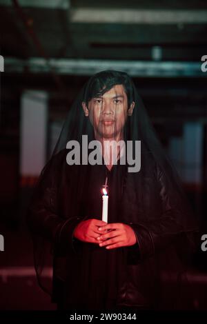 un adoratore del diavolo maschio con un velo trasparente sta eseguendo un rituale spettrale tenendo una candela in mano la notte Foto Stock